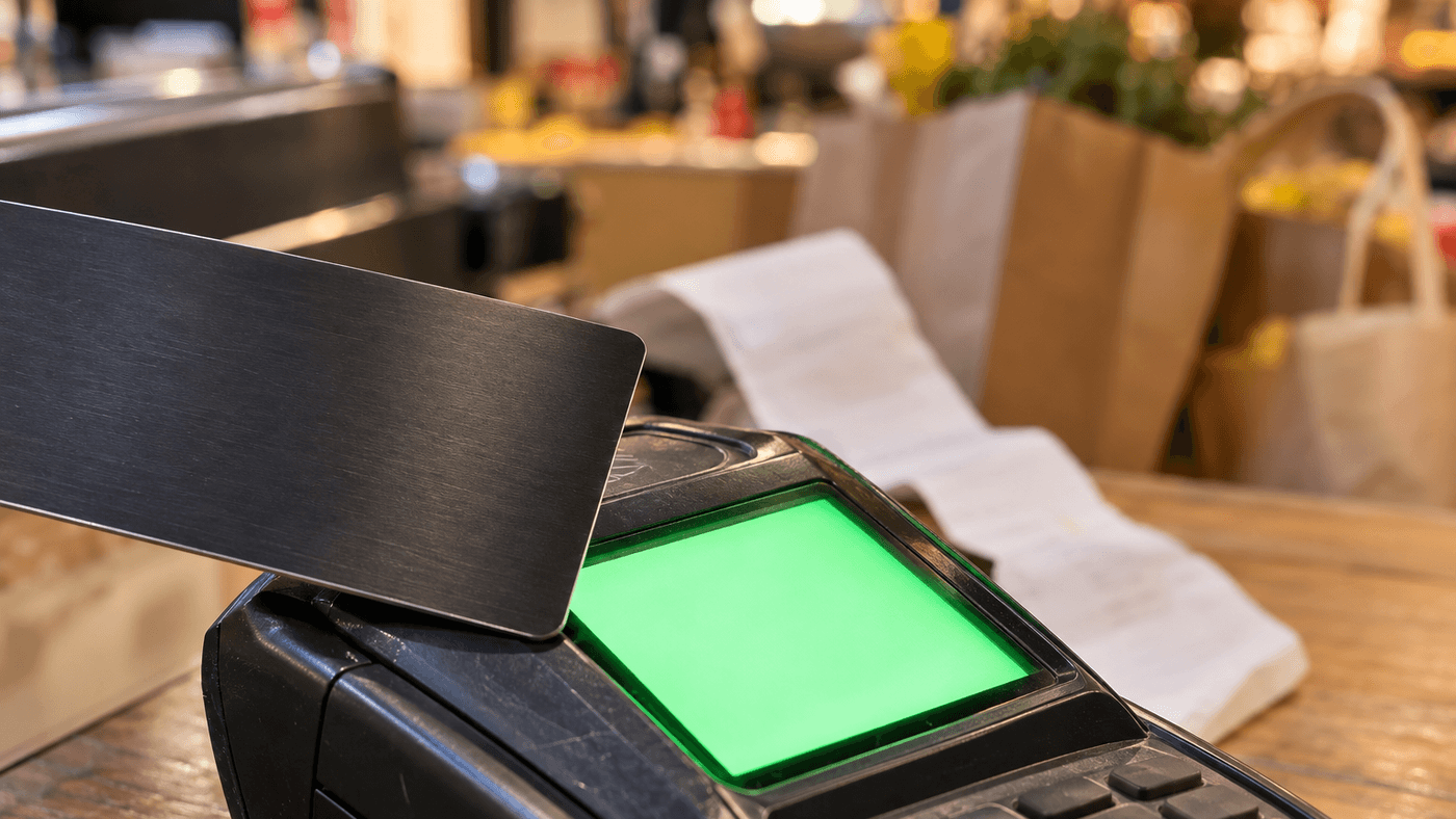 A blank dark payment card tapping a countertop card reader with a glowing green screen, shopping bags blurred behind