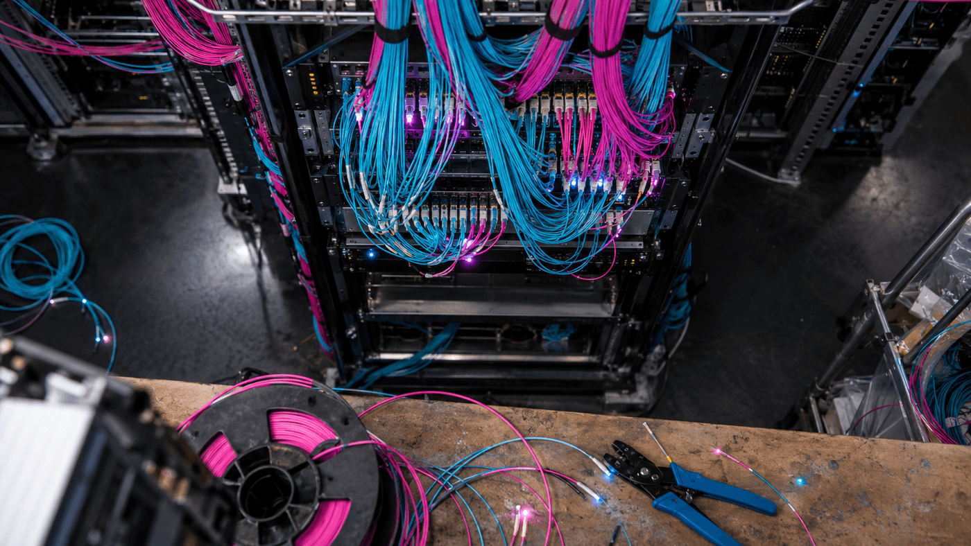 Blue and pink network cables patched into a rack of networking equipment, with cable spools and crimping pliers on a workbench in front