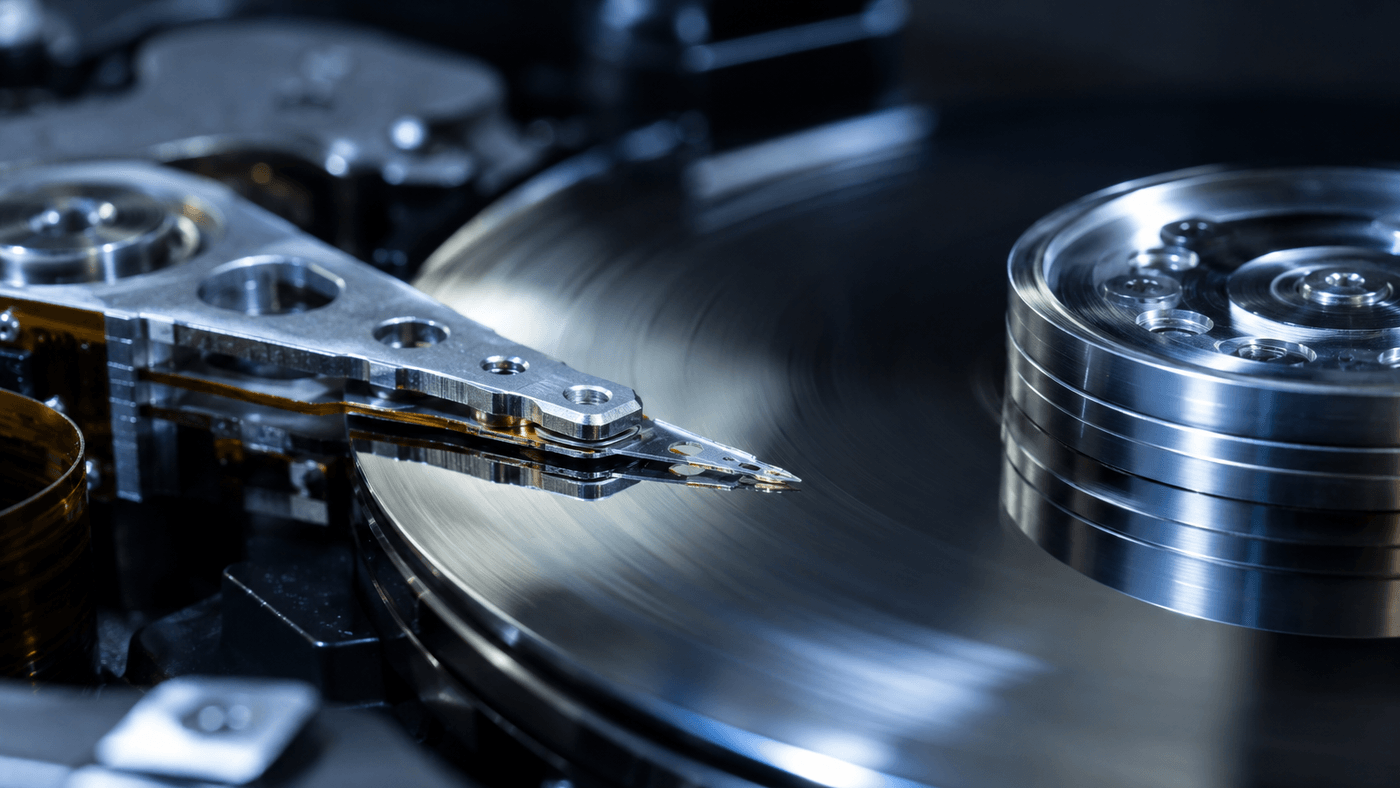 An opened hard disk drive seen up close, its mirror bright platter spinning under the read and write head arm in cool blue light